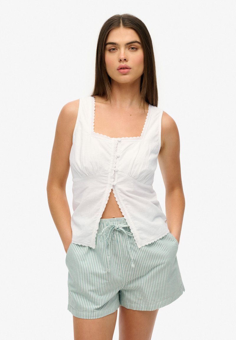 Vrouw in een witte mouwloze blouse met knopen en geschulpte randen, en lichtblauwe gestreepte shorts met een trekkoord, staand met haar handen in de zakken.