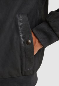 Zwarte leren jas met een patchzak, geribde manchetten en matte hardware met een logodetail. Soepele textuur en aansluitend ontwerp.