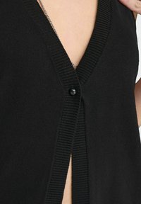 Cardigan nero a maglia con scollo a V, caratterizzato da un singolo bottone nero e dettagli a coste lungo i bordi. La texture appare morbida.