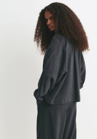 Femme aux longs cheveux bouclés portant une chemise et un pantalon gris foncé amples, se tenant de côté contre un fond clair et uni.