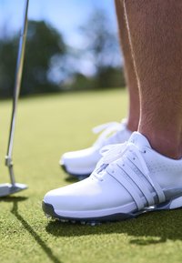 Bílé golfové boty s hladkým koženým povrchem, šněrovacím designem a perforacemi na boku. Umístěné na zelené trávě vedle putteru.