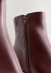 Burgundy nilkkurit, jotka on valmistettu sileästä synteettisestä materiaalista. Varustettu sivuvetoketjulla ja sauman suunnittelulla, joissa on kontrastivärisiä ompeleita.