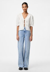 Top alb cu mâneci scurte și legătură în față, asortat cu blugi largi de culoare albastru deschis. Ținuta are un fit casual, lejer și un material texturat.