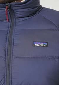 Doudoune bleu marine avec col montant, dotée d'une fermeture éclair et d'un patch logo Patagonia sur le côté gauche. Texture lisse.