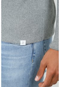 Maglione grigio lavorato a maglia con un motivo texturizzato, dotato di un'etichetta bianca sull'orlo, abbinato a jeans in denim blu.