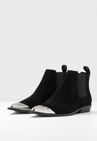 Botas Chelsea de ante negro con punta metálica brillante, paneles laterales elásticos y un tacón bajo y plano. Parte superior texturizada y diseño elegante.