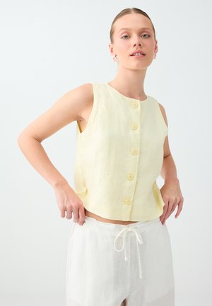 Giovane donna che indossa un top senza maniche giallo pallido con bottoni e pantaloni bianchi con coulisse su uno sfondo chiaro e uniforme.