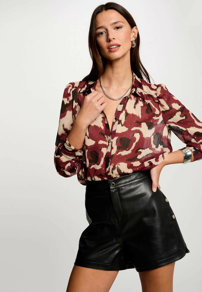 Blusa stampata rossa e beige con maniche a sbuffo e scollatura a V, abbinata a pantaloni corti in pelle nera a vita alta con dettagli a bottoni.