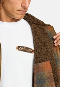 Bruine fleece jas met een gestructureerde binnenkant, geruite accenten in oranje en blauw, gedragen over een wit T-shirt met een zwart en oranje logo.