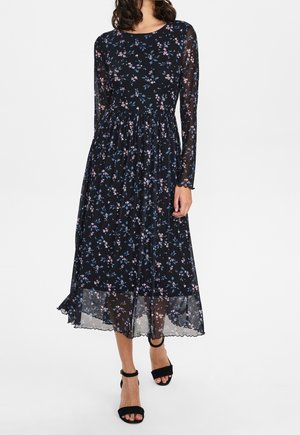 Kobieta ubrana w czarną sukienkę midi z długimi przezroczystymi rękawami i małym różowo-niebieskim kwiatowym wzorem, w połączeniu z czarnymi butami na obcasie z paskiem wokół kostki.