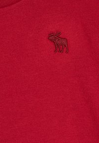 Tessuto di maglia rosso, con una sottile texture e un design di alce ricamato in una tonalità più scura, posizionato sul lato sinistro del petto.