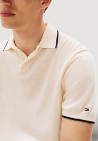 Polo beige con bordo del colletto blu navy, maniche corte e una piccola toppa con logo sulla manica. Tessuto morbido e design classico.