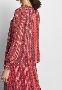 Rot gemusterte Bluse mit hohem Kragen und langen Ärmeln. Sie verfügt über eine Mischung aus floralen und geometrischen Designs aus durchscheinendem, leichtem Stoff.
