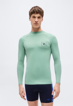 Mladý muž v svetlozelenom dlhom tričku na ochranu pred slnkom a čiernych plavkách s modrými a zelenými bočnými akcentmi stojaci pred jednoduchým pozadím.