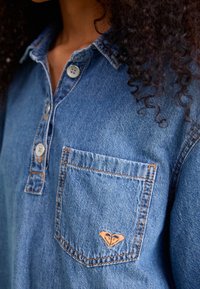 Chemise en denim bleu clair, dotée d'un col, de trois boutons et d'une poche poitrine avec un logo en forme de cœur. Texture douce et coutures visibles.