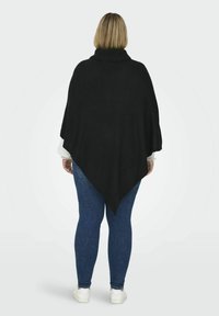 Poncho en tricot noir avec un col roulé haut et une forme triangulaire, superposé à une chemise blanche à manches longues et un jean bleu, avec une finition texturée.