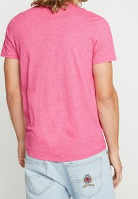 Tričko s krátkým rukávem a kulatým výstřihem vyrobené z hebkého růžového melírovaného materiálu, které má volný střih a hladkou texturu. K tomu jsou kombinovány světle modré džíny šortky.