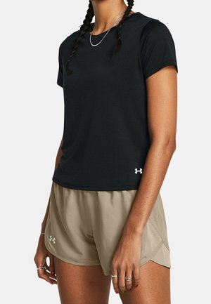 Person trägt ein schwarzes, kurzärmeliges Sportshirt und beige Laufshorts, steht mit entspannten Armen an den Seiten vor einem weißen Hintergrund.