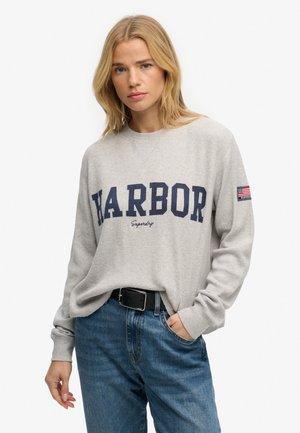 Lyshåret kvinde med skulderlangt hår iført en lysegrå "HARBOR" sweatshirt og højtaljede blå jeans, hånd i lommen, mod hvid baggrund.
