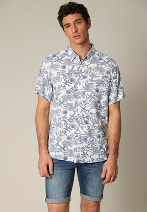 Chemise à manches courtes à boutons blanche avec un motif de feuilles de palmier bleues. Associée à un short en denim. Le tissu est léger et texturé.