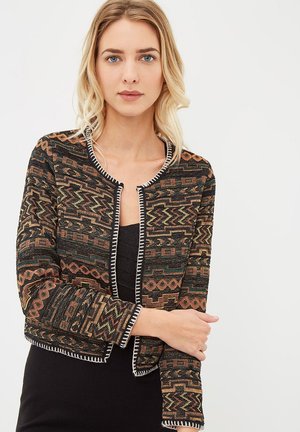 Una mujer con cabello rubio lleva un vestido negro y una chaqueta corta con intrincados patrones geométricos en naranja, verde y beige, y un ribete blanco.