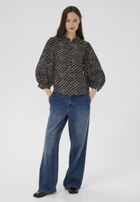 Fekete és bézs csíkos, puffos ujjú blúzt, bő szabású kék farmernadrágot és fekete cipőt viselő nő, aki a zsebében tartja a kezét.