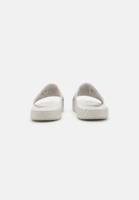 Fehér gumipadlós papucs vastag talppal és nyitott orral. A pántok minimalista, V alakúak, amelyek egyszerűsített esztétikát biztosítanak.