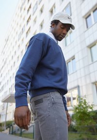 Donkerblauwe sweatshirt met geribde manchetten, gedragen met grijze jeans met een textuurpatch op de achterzak. Buitenshuis in een stedelijke omgeving.