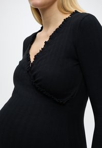 Κοντινό πλάνο ενός ατόμου που φοράει μαύρη ριμπ στρωτή κορυφή με βολάν στις άκρες και μακριά μανίκια, με φόντο απλό.