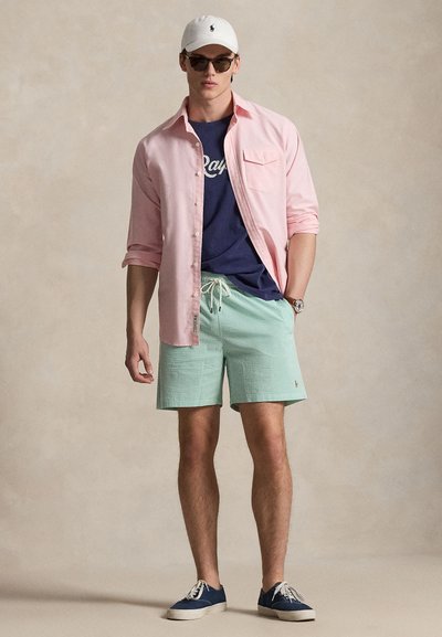 Ελαφρώς ροζ πουκάμισο με κουμπιά πάνω από ένα μπλε σκούρο t-shirt, σορτς με ριγέ μέντα, λευκό καπέλο, γυαλιά ηλίου και σκούρα αθλητικά παπούτσια, στέκεται σε ουδέτερο φόντο.