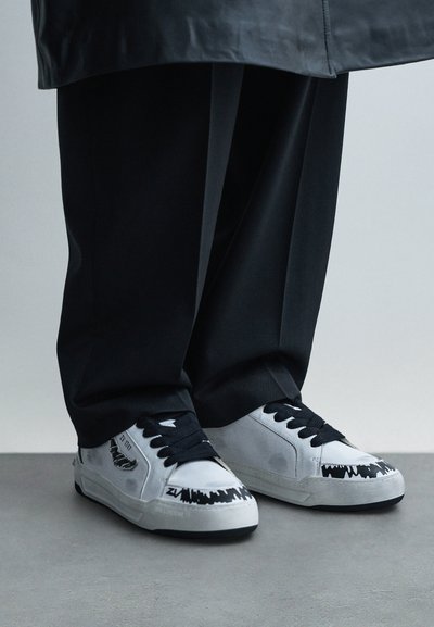 Sneakers in pelle bianca con dettagli neri e design grafico frastagliato sulla punta. Lacci neri e suola in gomma. Si abbinano a pantaloni neri.