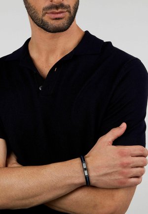 Uomo con polo nera a maniche corte, braccia incrociate, che mostra un braccialetto nero intrecciato con una chiusura in argento al polso.