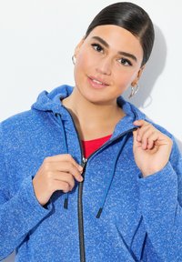 Blå fleece zip-up hoodie med en prickig textur, justerbar dragsko och svart dragkedja. Bärs över en röd topp.
