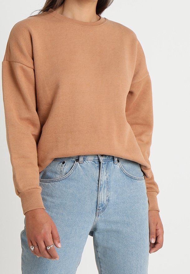 Femme portant un sweat-shirt uni marron clair à manches longues rentré dans un jean taille haute bleu clair, avec des bagues à la main gauche.
