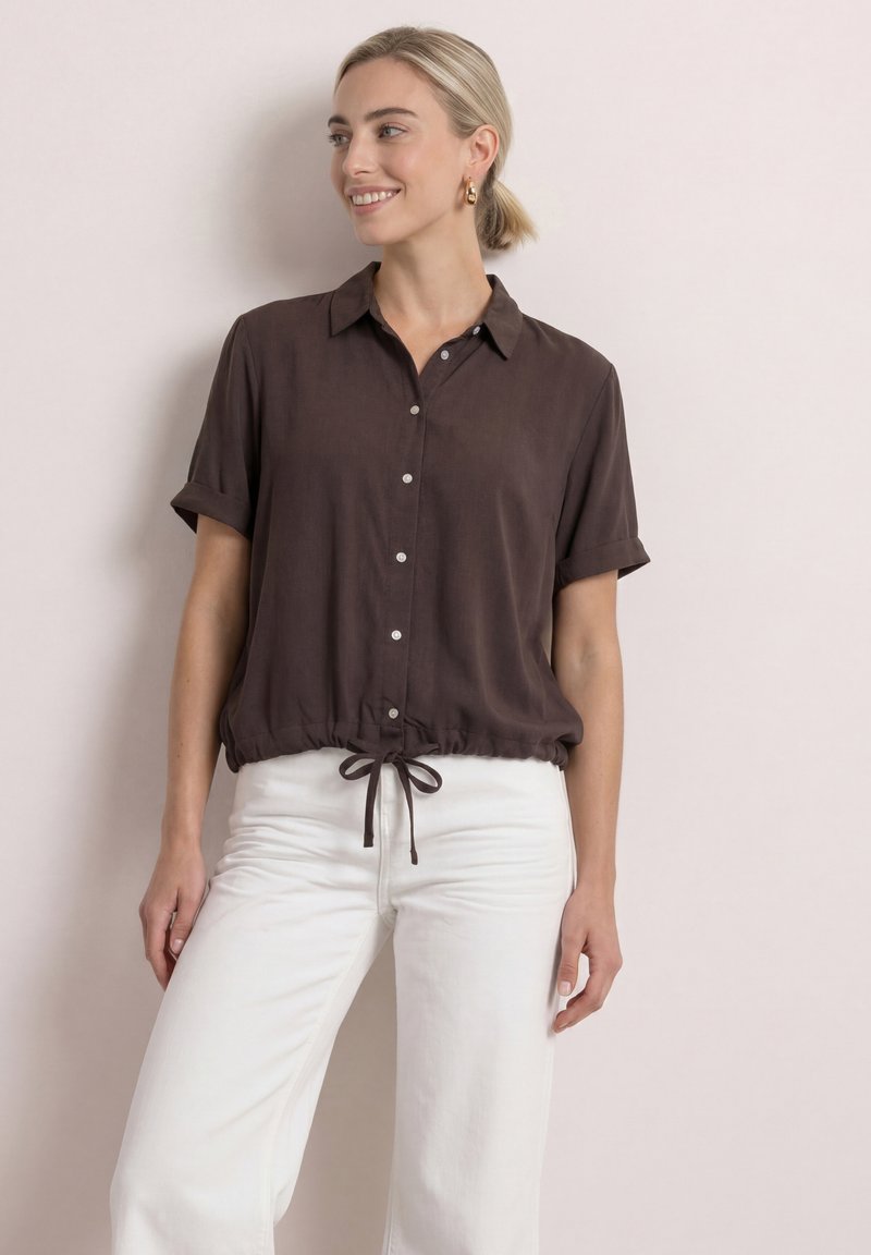 Femme aux cheveux blonds en queue de cheval basse porte une chemise marron foncé à manches courtes boutonnée avec un ourlet à cordon et un pantalon blanc, souriante de côté.