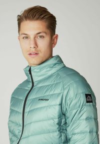 Svijetloplavi puffer jakna s visokim ovratnikom, horizontalnim šavovima, crnim patentnim zatvaračem i logotipom na lijevom rukavu.