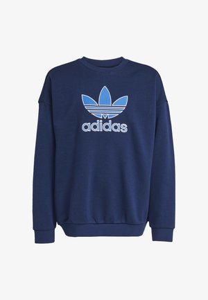 Adolescente con capelli ricci che cammina indossando una felpa Adidas blu navy e pantaloni della tuta abbinati con sneakers bianche.