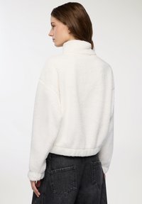 Giovane donna che indossa un maglione bianco soffice a maniche lunghe con colletto alto e jeans in denim neri, di profilo con la testa girata a sinistra.