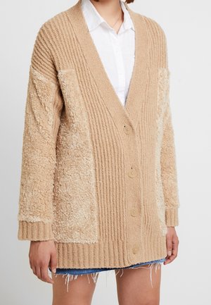 Frau trägt eine beige, geknöpfte Strickjacke mit gemischten Strick- und Flauschtexturen über einem weißen Hemd mit Kragen und einem Jeansrock mit ausgefranstem Saum.