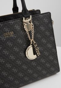Sac à main noir Guess avec motif monogramme, logo doré et breloque chaîne dorée ornée de pendentifs circulaires et rectangulaires Guess.