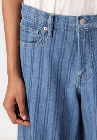 Jeans in denim azzurro chiaro a vita alta presentano strisce verticali, design a cinque tasche e una chiusura con bottone impreziosita da cuciture dorate.