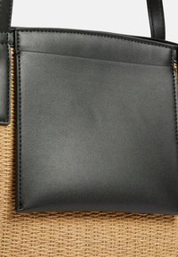 Poche en similicuir noir attachée à un sac en tissu tissé texturé de couleur marron clair, avec des coutures visibles le long des bords.