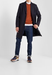 Navy coat over rust sweater and white shirt, paired with blue jeans and multi-colored trainers featuring dark and light accents and a textured finish.