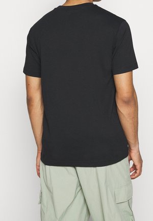 Personne portant un t-shirt noir uni à manches courtes et un short cargo vert clair, vue de dos sur fond blanc.