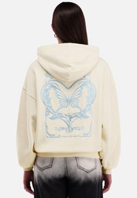 Geknipt geel sweatshirt met capuchon en een blauwe vlinderafbeelding op de achterkant. Zacht materiaal met elastische manchetten en een losse pasvorm.