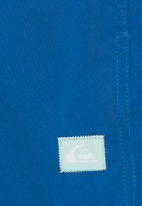 Ryški mėlynas audinys su siuvinėta etikete, ant šviesiai mėlyno fono esanti balta bangos logo. Lygus tekstūros, minimalus dizainas.