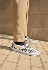 Sort-hvide ternede slip-on sneakers med hvid gummisål, iført med khakifarvede bukser og sorte sokker på en tekstureret belægning.