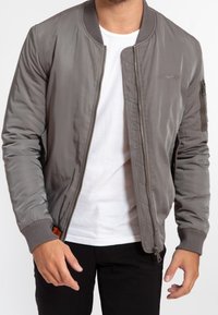 Man wearing unzipped gray bomber jacket over white t-shirt and black pants, standing with arms relaxed at sides.