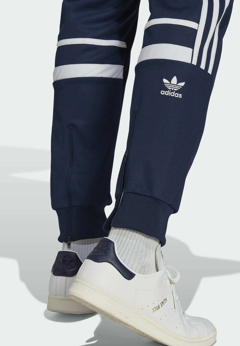Persona con pantalones de chándal negros de Adidas con rayas blancas y zapatillas Stan Smith blancas de Adidas con acentos en navy y calcetines blancos con canalé.
