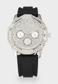 Reloj Guess que presenta una caja octagonal en tono plateado, acentos de cristales brillantes, una esfera plateada con dos subesferas y una correa de silicona negra.